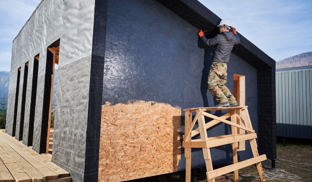 Male painter using paint roller, doing exterior paint work in a black color. Man worker building wooden frame house. Carpentry and construction concept.