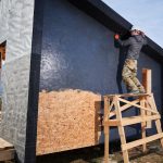 Male painter using paint roller, doing exterior paint work in a black color. Man worker building wooden frame house. Carpentry and construction concept.