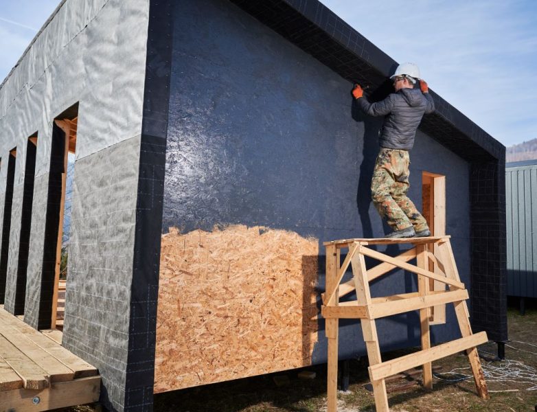 Male painter using paint roller, doing exterior paint work in a black color. Man worker building wooden frame house. Carpentry and construction concept.