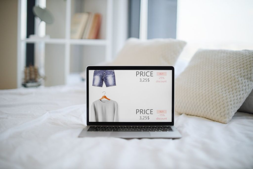 Modern wireless laptop with online apparel shop on screen placed on comfy bed with white linen. Discount of 15 percent and 25 percent for online order. Consumerism concept.