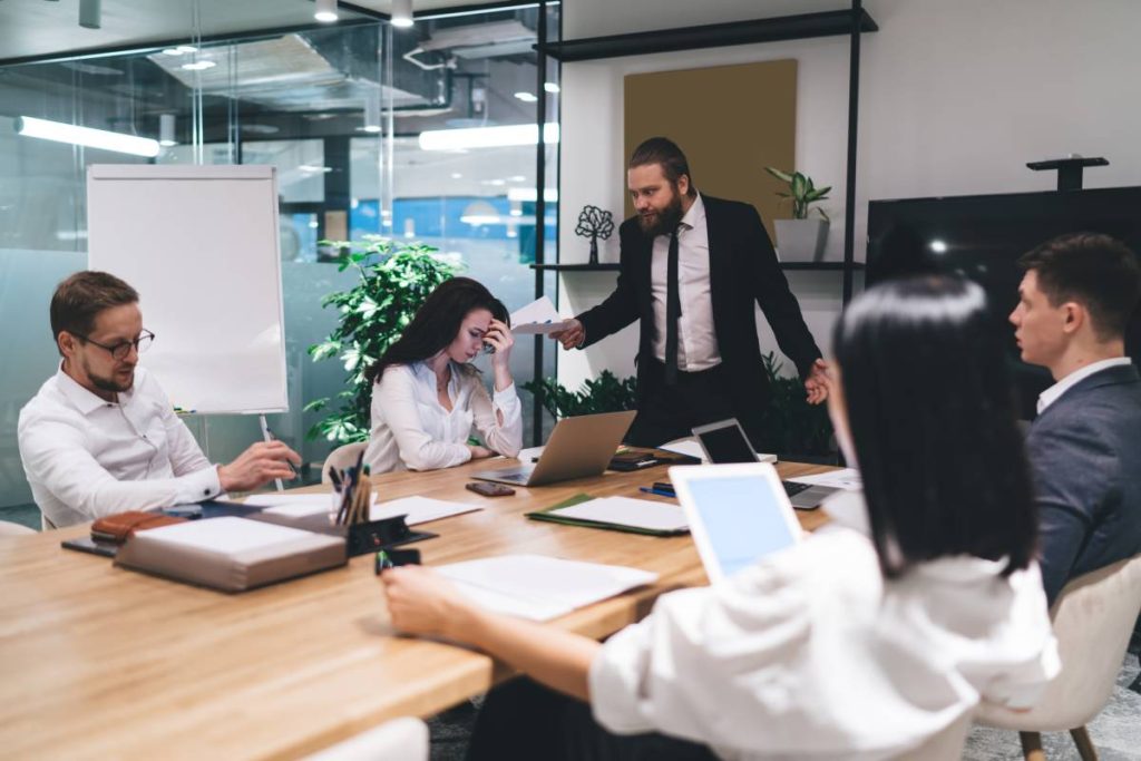 Dissatisfied male boss in suit scolding frustrated female worker sitting at table with colleagues during business meeting in modern workplace