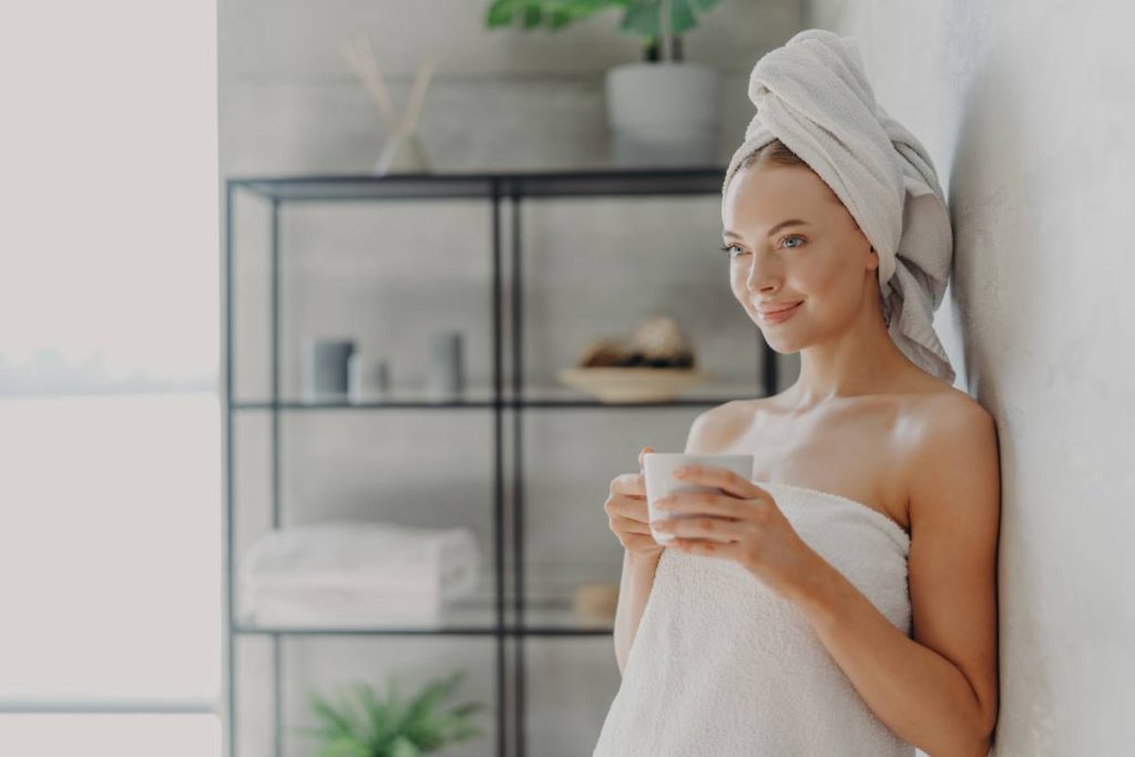 Horizontal shot of beautiful young woman with healthy skin wrapped in bath towel, poses with mug of tea, concentrated into distance, poses near wall over home interior, enjoys cozy atmosphere