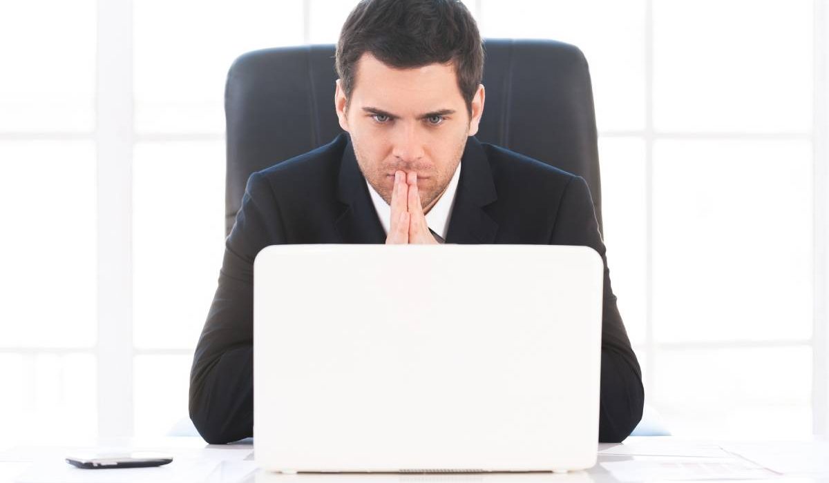 Finding out solution. Worried young man in formalwear looking at the computer and holding hands clasped while sitting at his working place