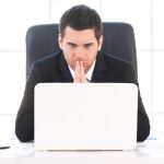 Finding out solution. Worried young man in formalwear looking at the computer and holding hands clasped while sitting at his working place