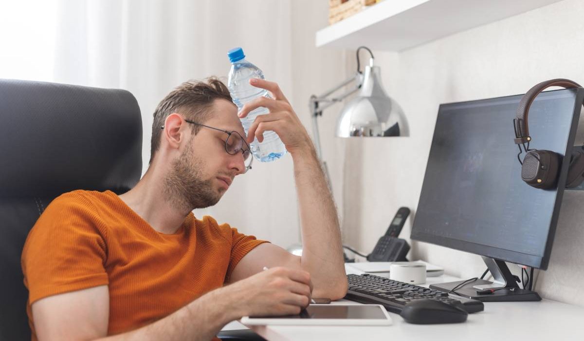 Working at home man suffering from heat and thirst cools down with water bottle at hot summer day. Heat weather without air conditioning