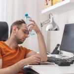 Working at home man suffering from heat and thirst cools down with water bottle at hot summer day. Heat weather without air conditioning