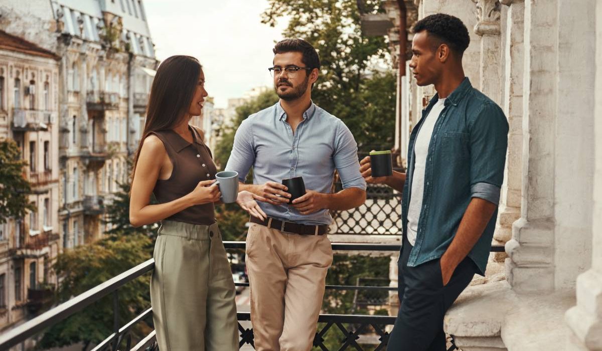 Conversation. Young cheerful colleagues in casual wear holding cups and talking with each other while standing on the balcony. Coworkers resting. Coffee break