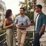 Conversation. Young cheerful colleagues in casual wear holding cups and talking with each other while standing on the balcony. Coworkers resting. Coffee break