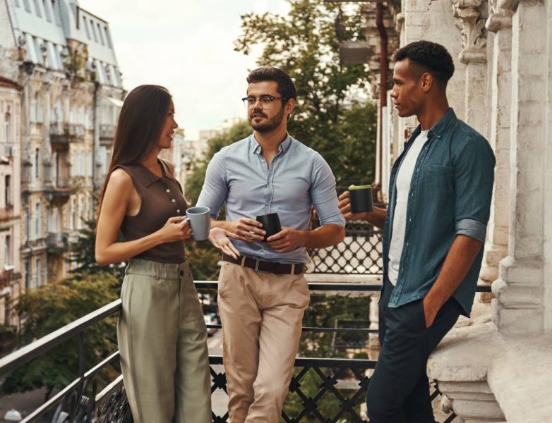 Conversation. Young cheerful colleagues in casual wear holding cups and talking with each other while standing on the balcony. Coworkers resting. Coffee break