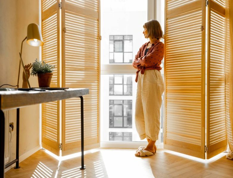 Young woman stands alone by the window blinds at cozy and sunny living room of modern apartment in beige tones. Quarantine, loneliness and life at home