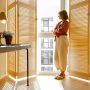Young woman stands alone by the window blinds at cozy and sunny living room of modern apartment in beige tones. Quarantine, loneliness and life at home