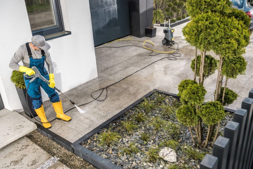 Caucasian Men Pressure Washing House Surrounding Concrete Elements. Cleaning Paths and Driveway Using Powerful Washer.