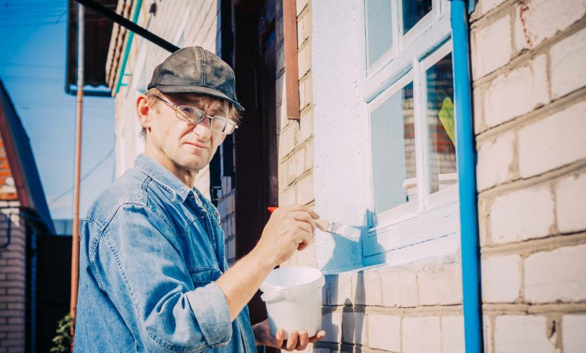 Caucasian Mature Man In Glasses And Cap Painting The Window Frame Outside Of The Private Brick House In Sunny Day.