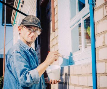 Caucasian Mature Man In Glasses And Cap Painting The Window Frame Outside Of The Private Brick House In Sunny Day.