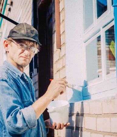 Caucasian Mature Man In Glasses And Cap Painting The Window Frame Outside Of The Private Brick House In Sunny Day.