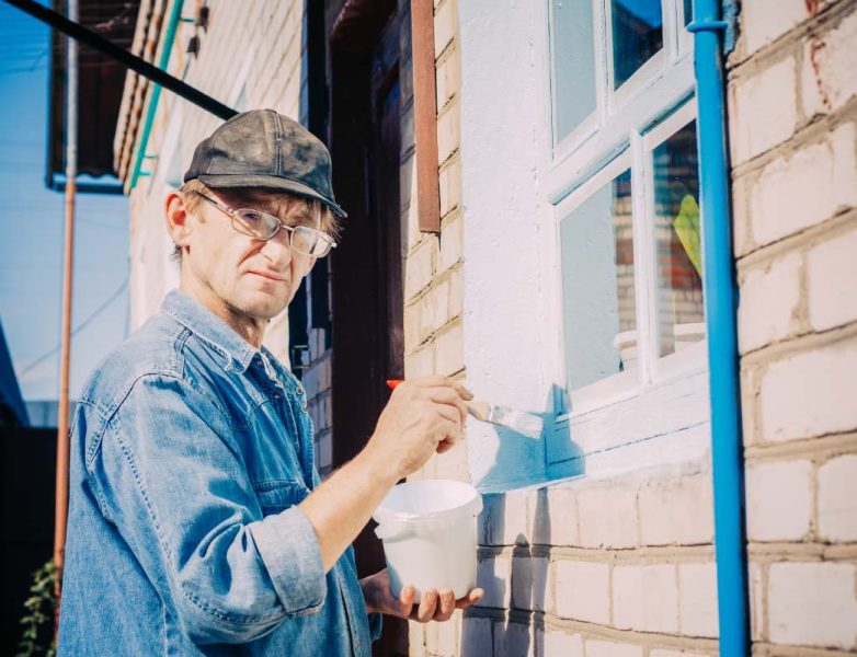 Caucasian Mature Man In Glasses And Cap Painting The Window Frame Outside Of The Private Brick House In Sunny Day.