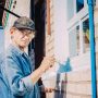 Caucasian Mature Man In Glasses And Cap Painting The Window Frame Outside Of The Private Brick House In Sunny Day.