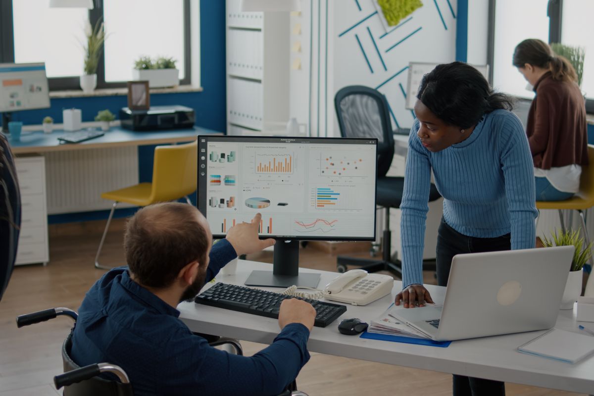 African bookkeeper working with handicapped invalid immobilized colleague calculating profit on laptop, taking notes on pc, analysing financial documents sitting at desk in start-up financial company.