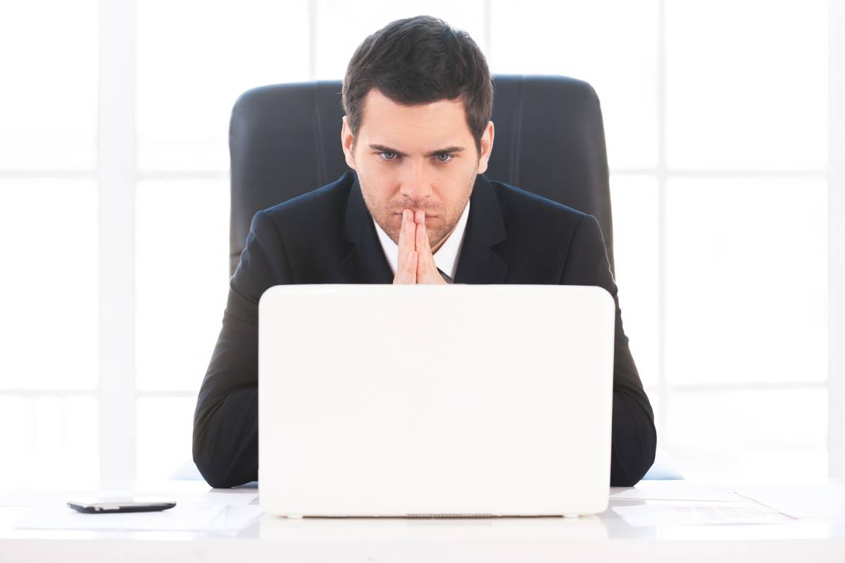 Finding out solution. Worried young man in formalwear looking at the computer and holding hands clasped while sitting at his working place