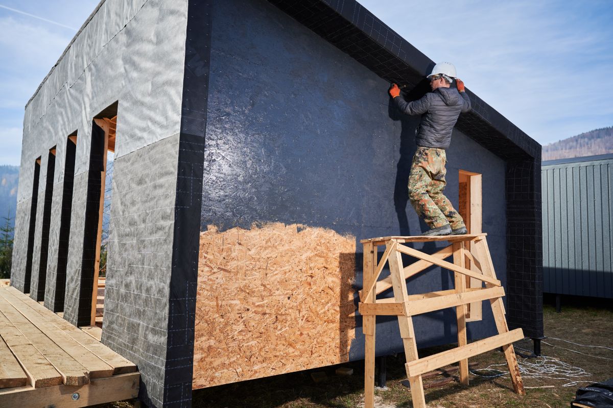 Male painter using paint roller, doing exterior paint work in a black color. Man worker building wooden frame house. Carpentry and construction concept.