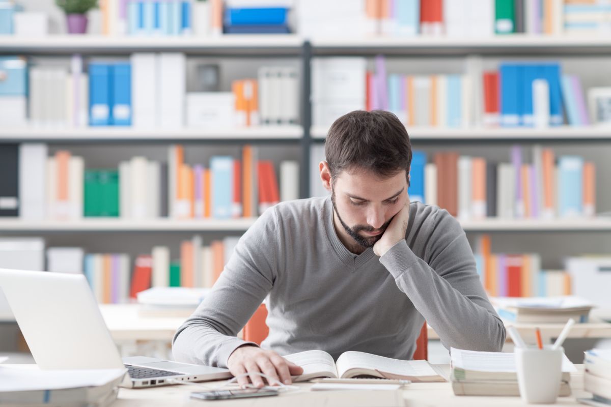 Young focused smart man in the office studying and reading a book, shelves on background