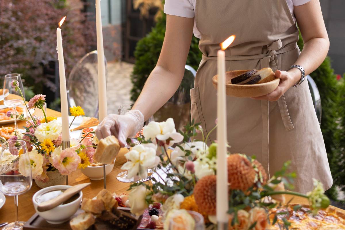 Wedding catering service. Banquet. Chairs and a table for guests, decorated with candles, are served with cutlery and crockery and covered with yellow tablecloth. Waiter setting table stands on a green lawn in the backyard banquet area